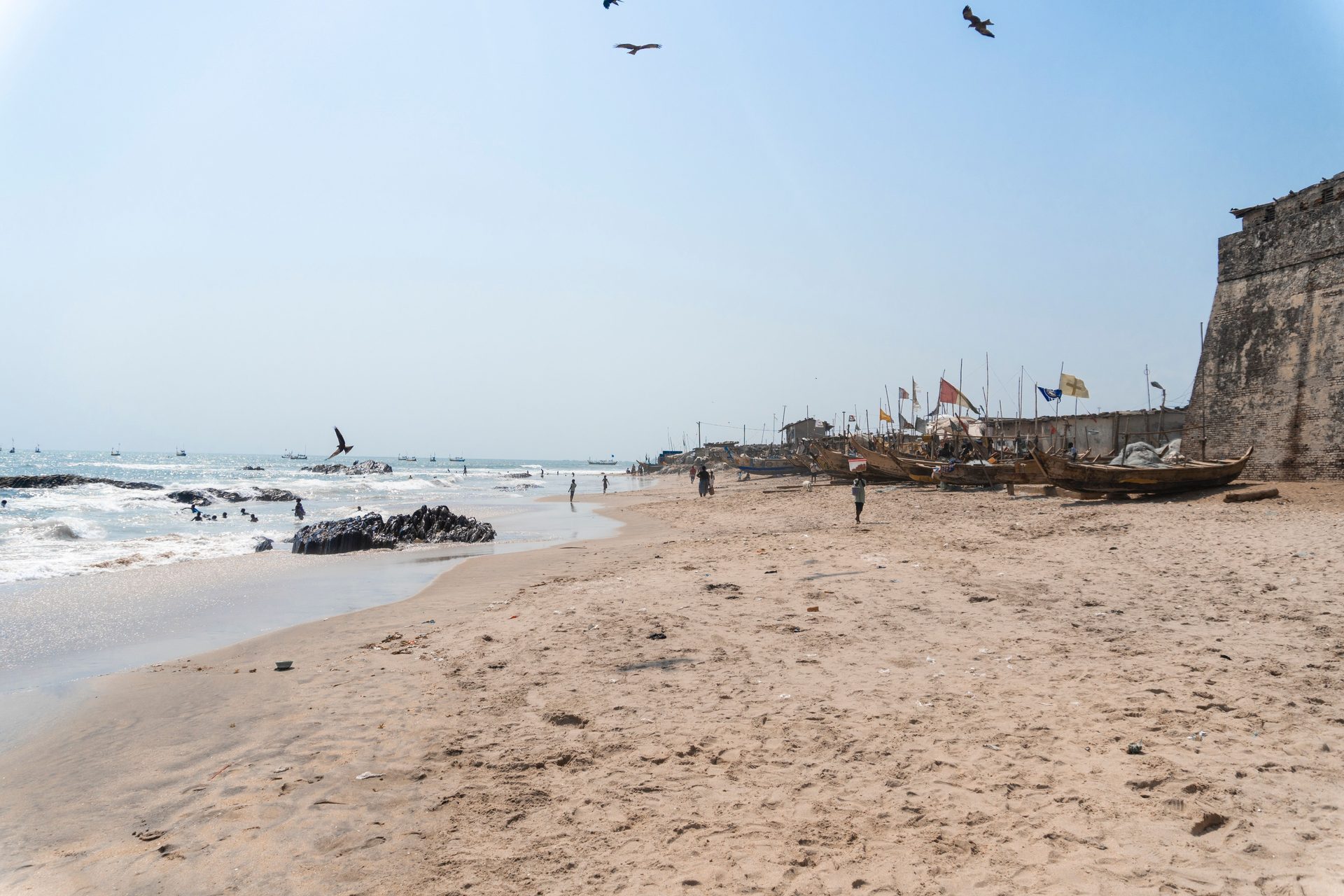 Fishing canoes and flags on a sandy Ghana beach beside the stone wall of an old coastal fort
