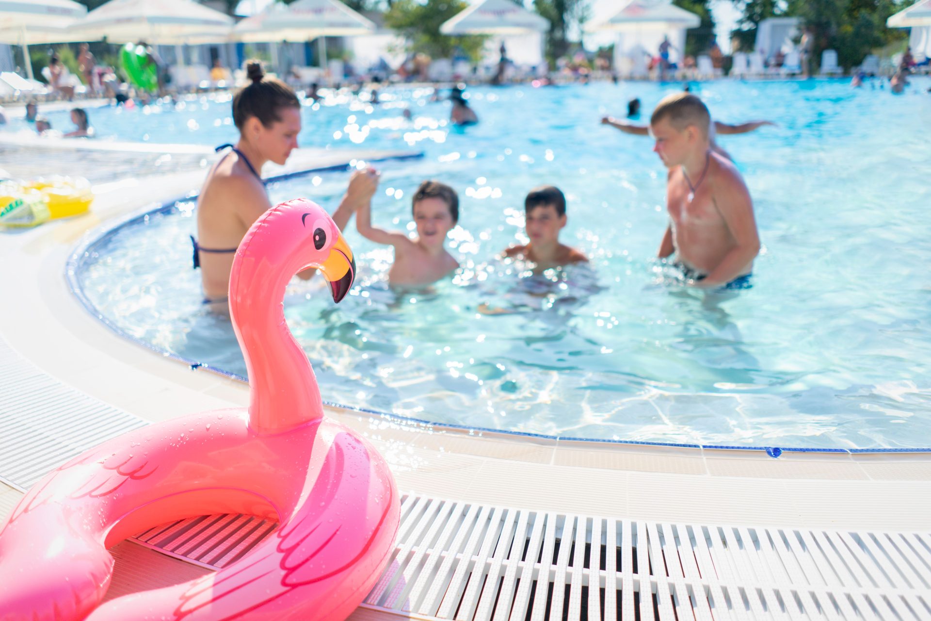 A woman and three children playing in a sunny resort swimming pool, with a pink flamingo float on the poolside