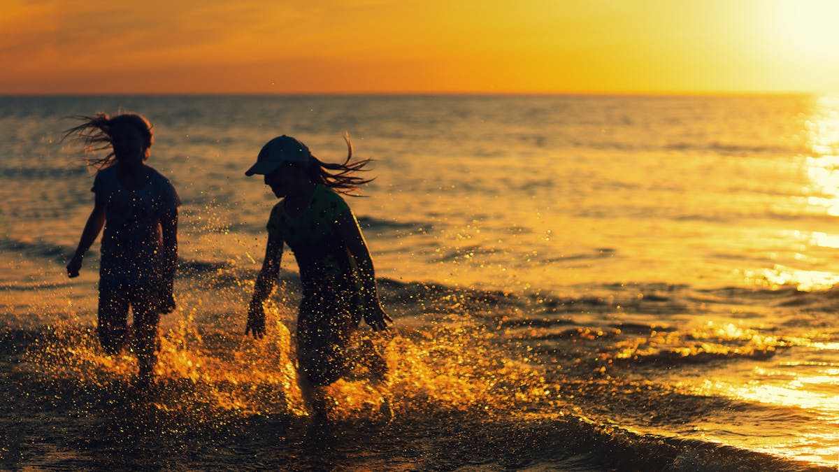 Two children joyfully splash in the water at a beach during sunset, capturing carefree summer fun