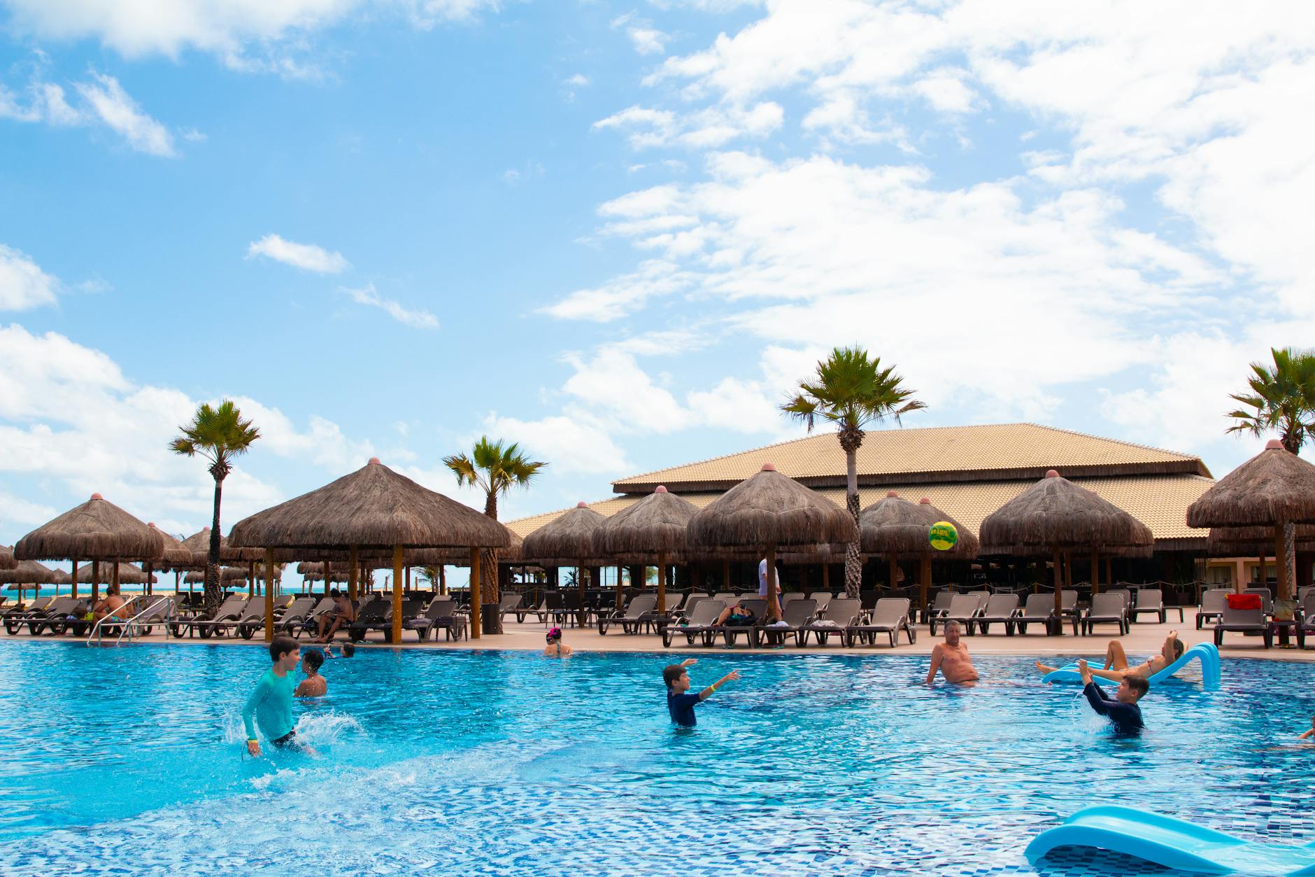 Families enjoying a sunny day at a resort pool with palm trees and thatched umbrellas &mdash; the all-inclusive resort context that defines most Cabo family vacations