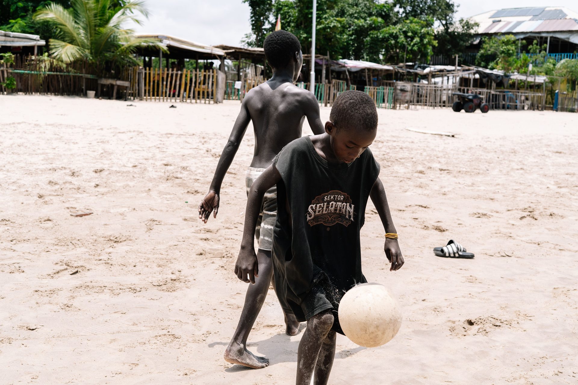 Two boys playing soccer on a sandy Lagos beach in front of palm-thatched beach huts