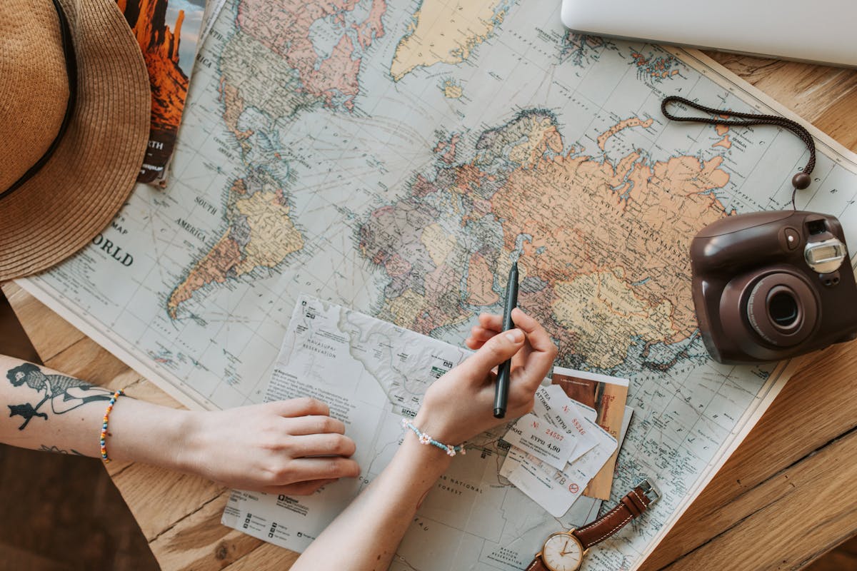 Hands planning a trip over a paper world map with tickets, a camera and a sun hat