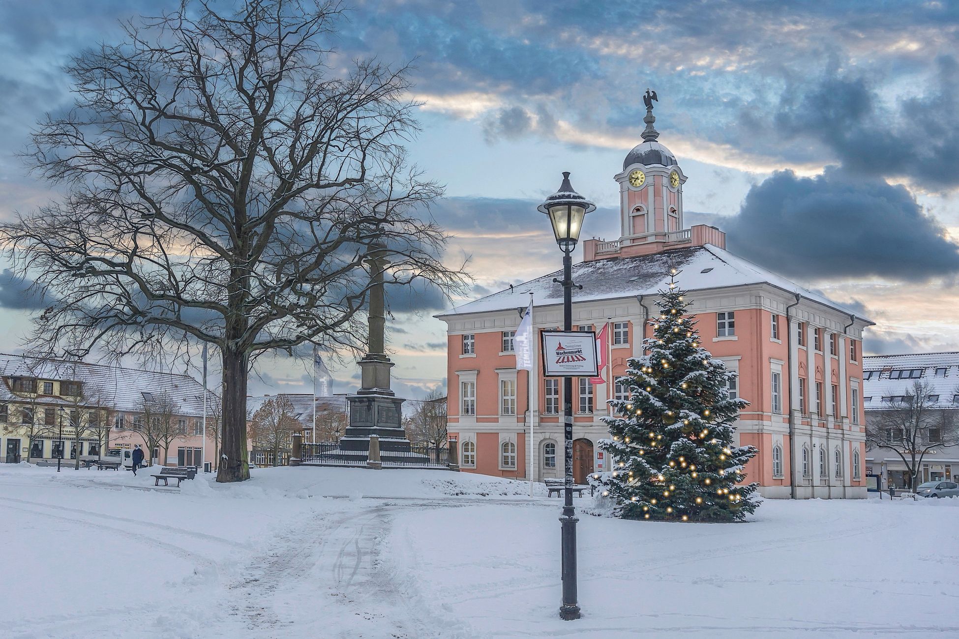 Snowy town square at dusk with a lit Christmas tree beside a historic clock-tower town hall