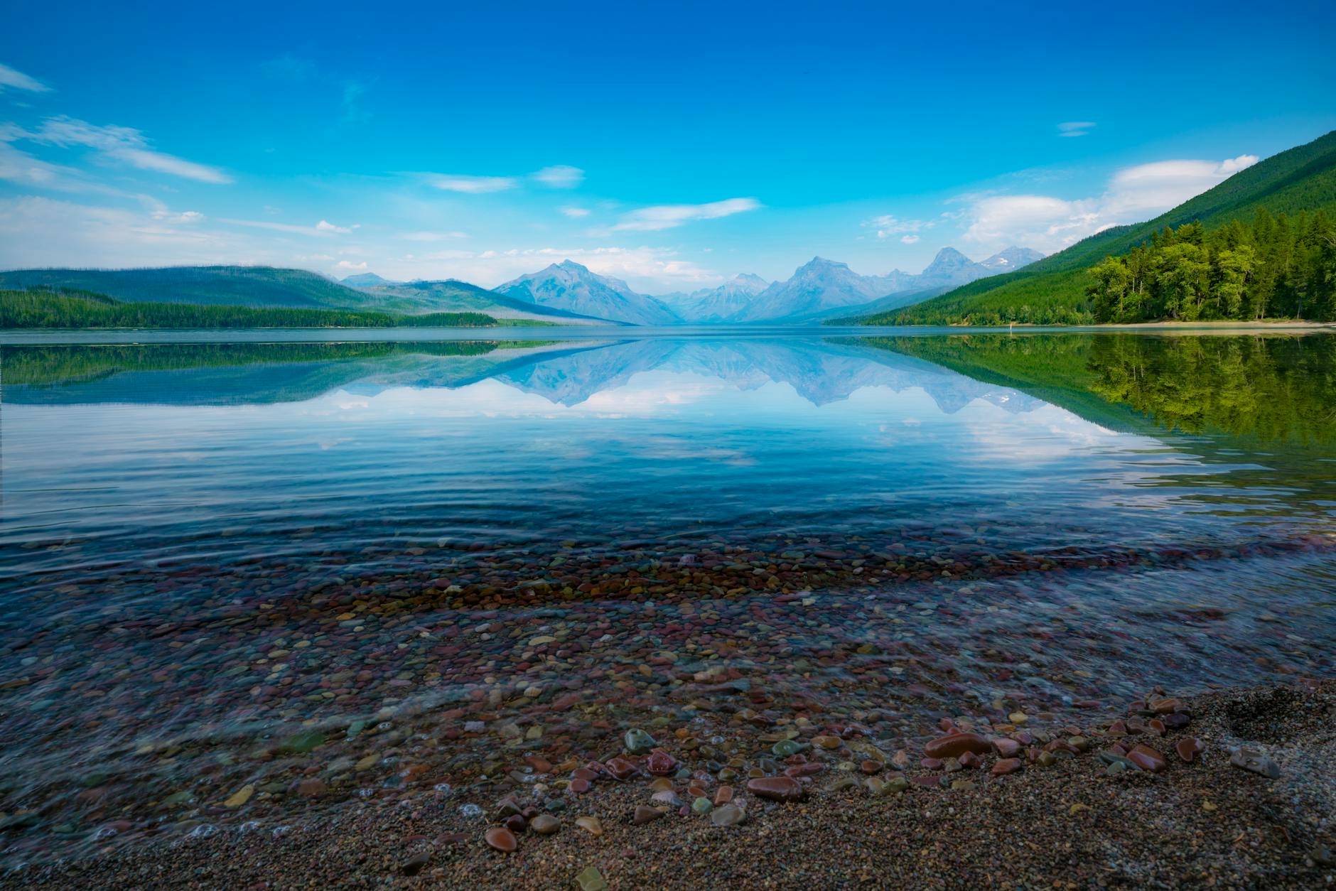 Clear water with colorful cobblestones at Lake McDonald with mountain backdrop in Glacier National Park