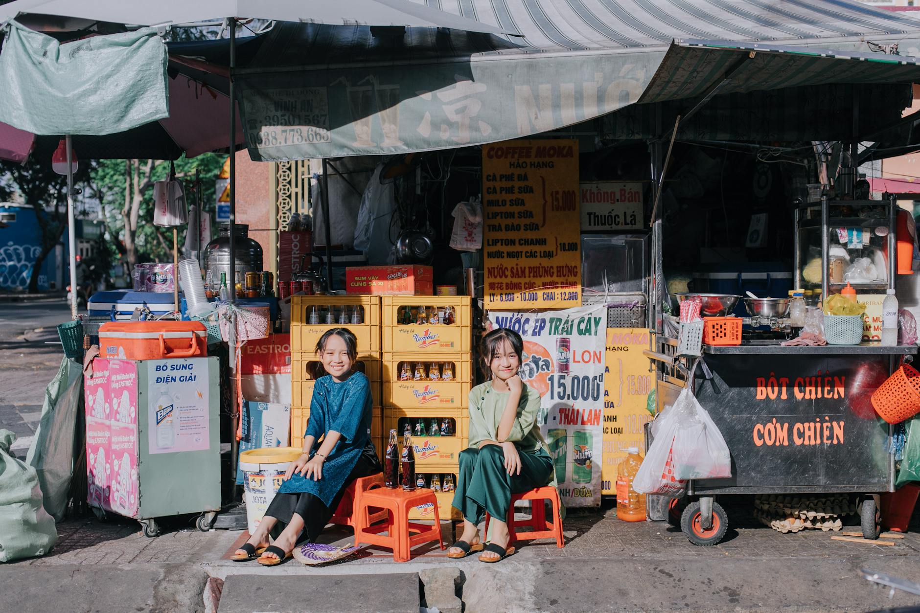 Two children sitting happily outside a colorful street market stall in Ho Chi Minh City