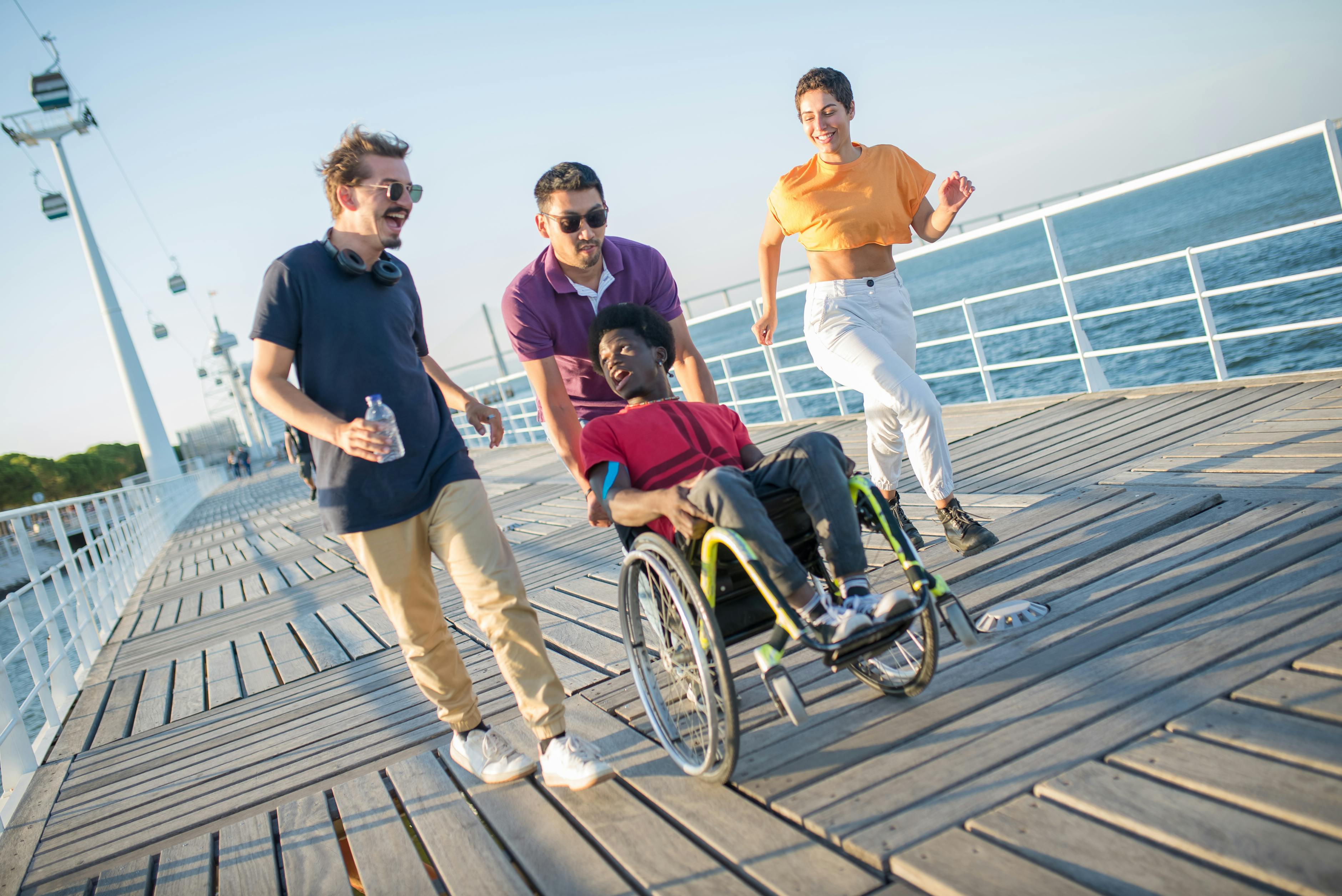 Friends on a sunny boardwalk pushing a wheelchair together &mdash; the social, family-of-friends side of an accessible vacation